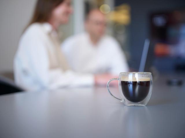 Billede af et glas med sort kaffe i og to mennesker, der sidder sløret i baggrunden ved en computer